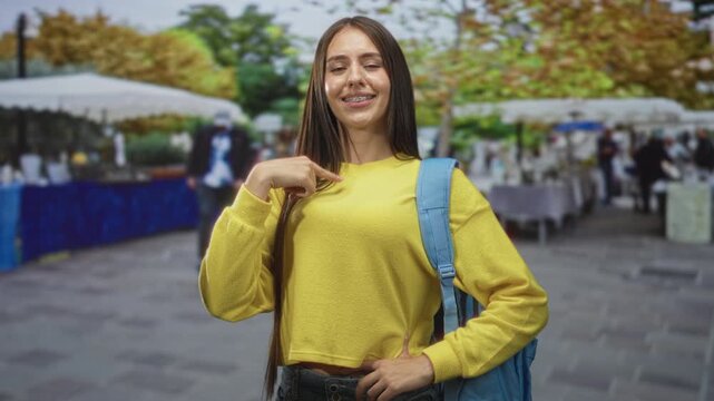 Teen girl student wearing yellow sweater and backpack points finger to chest at a bustling street market; youthful confidence.