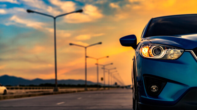 Blue compact SUV car open headlamp light parked on concrete road near the mountain at sunset with beautiful sky and clouds. Road trip travel. Automotive industry. Electric and hybrid car technology.