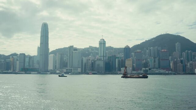 Wide static shot of Hong Kong Island skyline featuring Two IFC tower across Victoria Harbour, with tugboat and crane barge in foreground under overcast cloudy sky, cinematic daytime mood