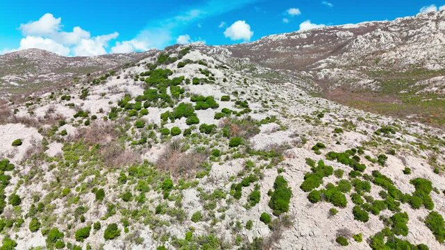 Drone pulling back near a Velebit peak in Zadar County, showing rocky slopes, scarce bushes, and the vast mountain landscape opening up in bright sunshine.
