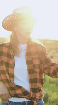 Middle-aged beautiful woman farmer in a hat has online conference with agribusiness partners standing in a field at sunset. Agricultural specialist video call by phone. Slow motion.