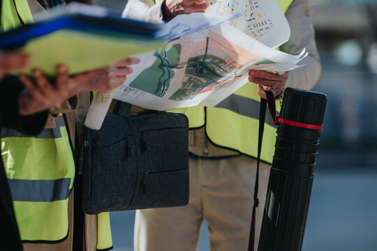 Construction engineers review site plans and maps while wearing high-visibility safety vests. Close-up of colleagues discussing project layouts and documents on a construction site.