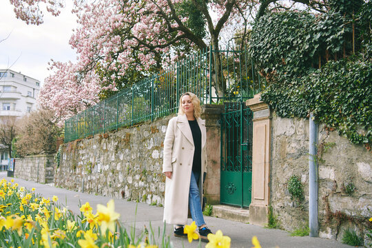 Mature woman walking along spring street with blooming flowers and magnolia trees, stylish 45 year old female enjoying calm urban life, peaceful outdoor scene in European town