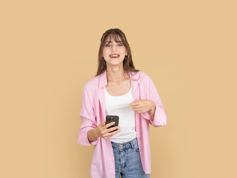 Smiling caucasian woman holding smartphone and pointing at it with inviting expression, promoting app or message concept image, wearing pink shirt and denim, isolated beige background with copy space.