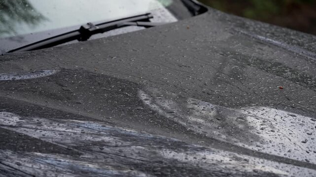 Close-up of raindrops on dark car hood during rainy weather. Water beads and streaks create reflective textured surface with windshield wiper in background. Moody outdoor scene, shallow depth of field