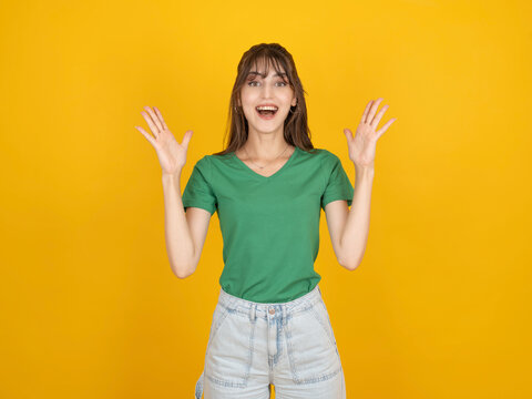 Excited caucasian woman raising hands with joyful surprised expression, celebrating success or good news, wearing green t shirt and denim, isolated on bright yellow studio background with copy space.