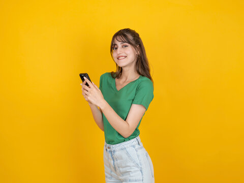 Smiling caucasian woman holding smartphone and typing message with cheerful expression, wearing green t shirt and denim, isolated on yellow studio background with copy space. Lifestyle concept.