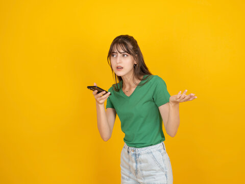 Confused caucasian woman holding smartphone and gesturing with questioning expression, reacting to unclear message, wearing green t shirt and denim, isolated yellow studio background with copy space.