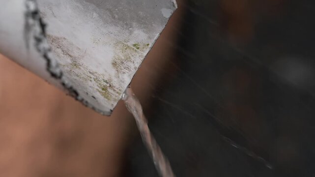 Close-up of rainwater flowing from drain pipe on house exterior. Water stream pouring from roof gutter downspout during rainy weather, outdoor drainage system detail with shallow depth of field