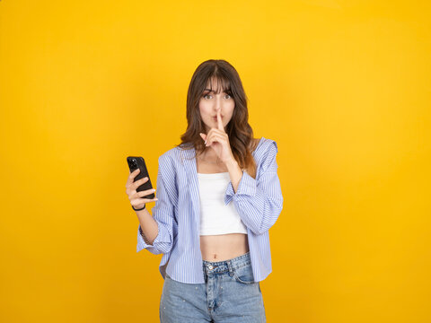 Caucasian woman holding smartphone and making silence gesture with finger on lips, expressing secrecy, wearing striped shirt and denim, isolated on bright yellow studio background with copy space.