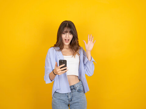 Shocked caucasian woman looking at smartphone with raised hand in surprise, reacting to unexpected message, wearing striped shirt and denim, isolated on yellow studio background with copy space.