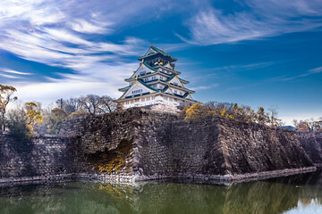 Obraz premium Magnificent view of Osaka Castle in spring, Japan. The historic fortress is surrounded by stone walls, a peaceful moat, and blooming cherry blossoms under a vibrant blue sky in early April.
