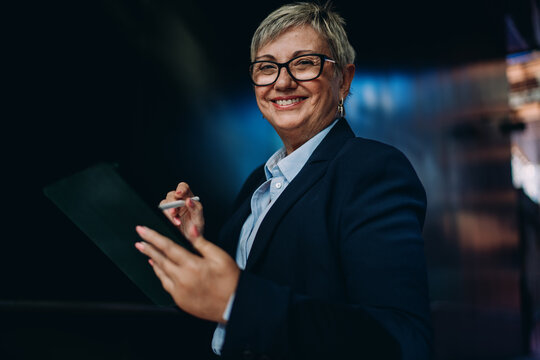 Mature businesswoman in formal suit holding stylus and tablet while looking back confidently, symbolizing digital control and awareness in a mobile-first workspace.