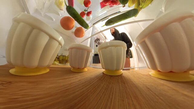 View from inside a refrigerator as a man reaches in to grab a grapefruit. The wide-angle perspective captures various foods on a wooden shelf, with a clear focus on the action