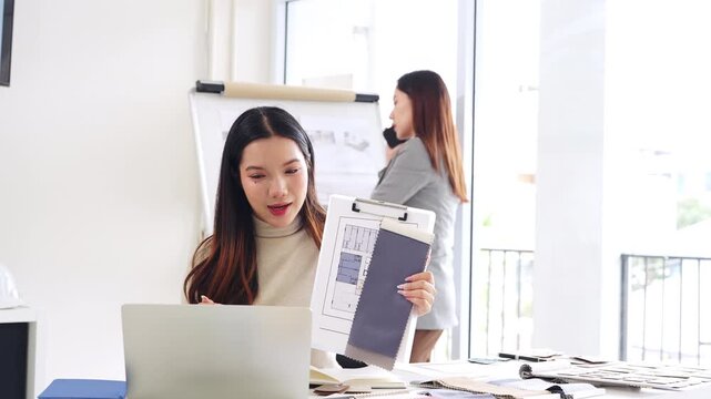 Asian Interior designer sitting in front of laptop talking with client on video call, showing architecture floor plan blueprint of residence and pantone. meeting and online conference.