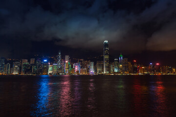 Obraz premium Panoramic night view of Hong Kong illuminated skyline along Victoria Harbour, featuring soaring skyscrapers, neon lights and vivid reflections on the water under a dramatic clouded sky