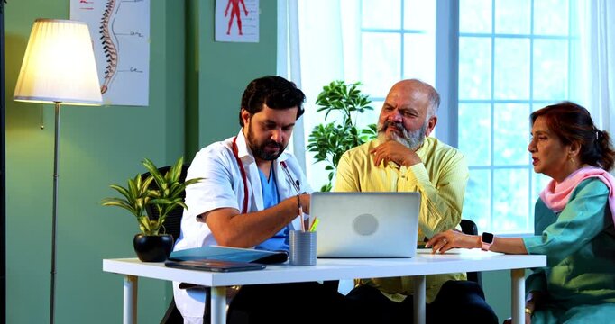 Professional Indian young male doctor using laptop computer with Asian senior retired couple patients across a table in a modern clinic; healthcare, digital technology, and old age care concept.