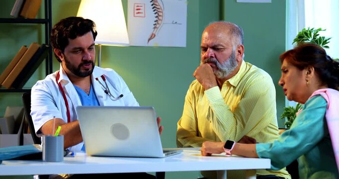 Professional Indian young male doctor using laptop computer with Asian senior retired couple patients across a table in a modern clinic; healthcare, digital technology, and old age care concept.