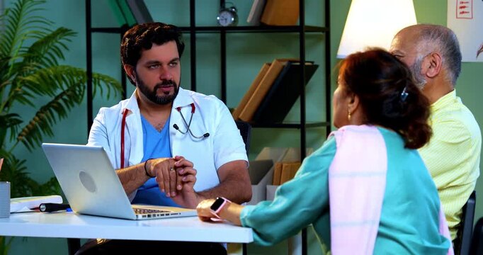 Professional Indian young male doctor using laptop computer with Asian senior retired couple patients across a table in a modern clinic; healthcare, digital technology, and old age care concept.