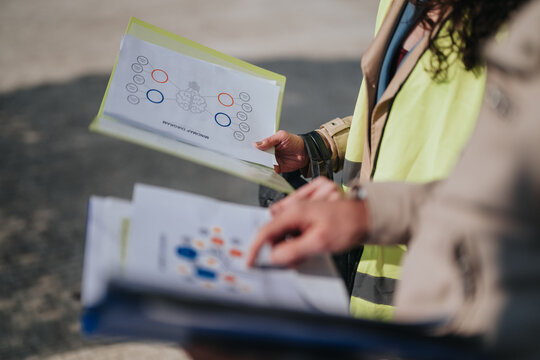 Two workers wearing high-visibility vests review printed diagrams and flowcharts held in folders. They examine project charts outdoors during a planning discussion.