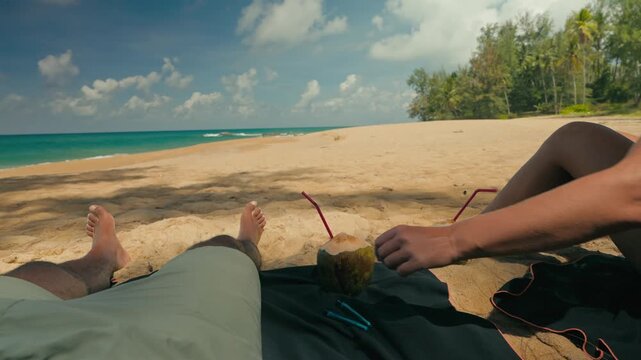 Close-up of a couple&rsquo;s legs relaxing on a tropical beach with coconuts and rolled items in a tube on towel. Sunny waves, sand, and palm trees create a peaceful, laid-back vacation atmosphere