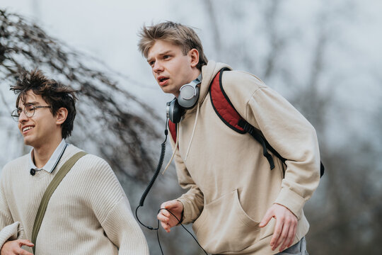 Two teenage friends walk together outdoors, one wearing headphones and a backpack. They appear carefree and chatty as they stroll on a blustery day.