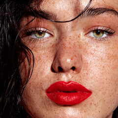 Naklejka premium Portrait of a young woman with bold red lipstick, tousled black hair, and freckles, looking straight at the camera against a dark gray background