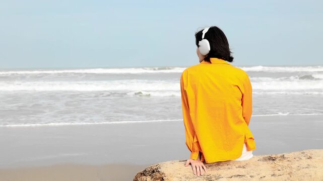 Static shot of person adjusting headphones while sitting on rock at sandy ocean beach during daytime, calm seascape view