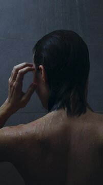 Slow static tracking shot of woman washing and rinsing hair in dim tiled shower bathroom back view with hands on head