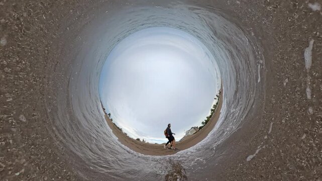 A couple walks along the shoreline as a wave rushes toward them, captured in a Tiny Planet effect that wraps the beach, water, and sky into a dynamic spherical panorama