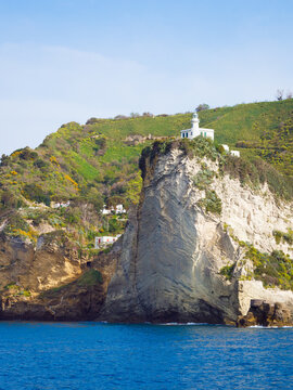 White lighthouse stands on top of vertical rock cliff above Mediterranean sea. Green hills and houses are in background under clear blue sky. Scenic coastal landscape of Phlegraean Fields region