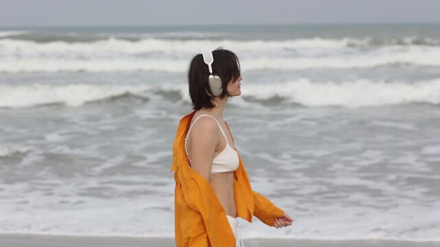 Tracking shot of woman walking along shoreline with headphones and orange jacket on sandy beach under overcast sky