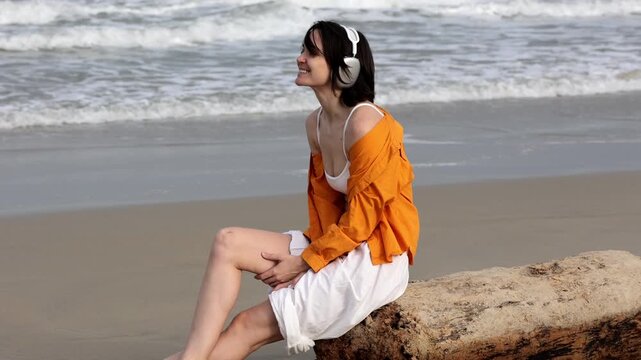 Panning tracking shot of woman wearing headphones turning and looking toward ocean while sitting on driftwood beach