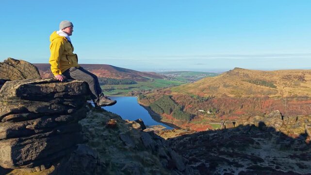 Drone POV of a man sitting on the rock with the panoramic view of Dovestone reservoir. Dove Stone Reservoir lies at the convergence of the valleys of the Peak District National Park, Greenfield, UK.
