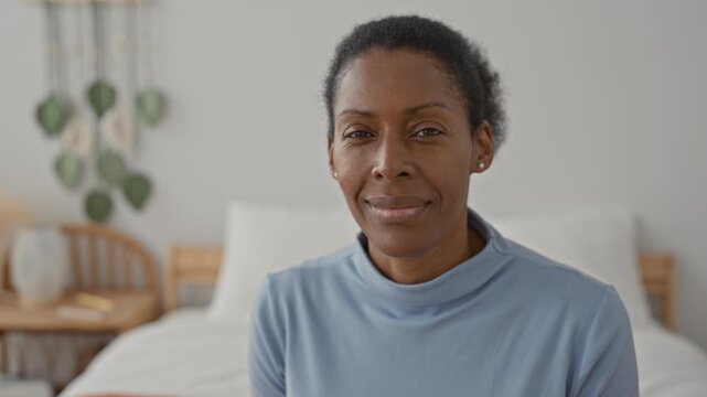 Woman smiling on bed in a residential building, wearing light blue turtleneck and stud earring while looking at camera; contentment.