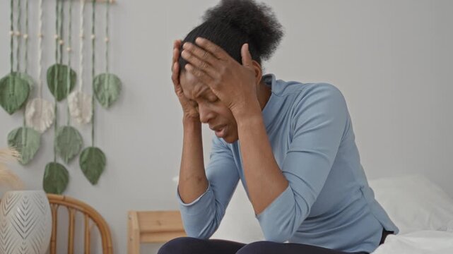 Woman holding head with both hands while seated on a bed in a building, eyes closed and grimacing with a visible headache and tense posture; distress.