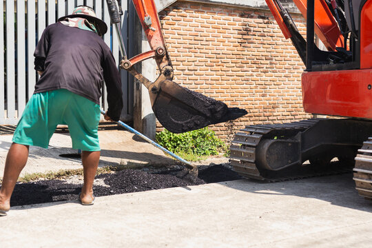 A worker using a rake to level fresh asphalt alongside an excavator during road repairs.