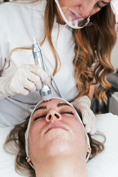 A professional aesthetician in a white uniform uses a radiofrequency skin tightening device on a woman's forehead during a rejuvenating facial procedure in a wellness spa clinic.