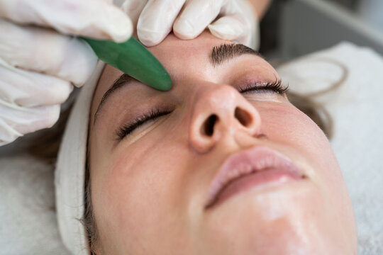 A professional esthetician wearing gloves uses a green jade gua sha stone to massage the forehead of a relaxed female client during a skin rejuvenation treatment at a wellness clinic.