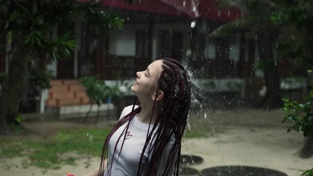 Young lady enjoying rainy day antics. Youthful woman shakes water from her braided hair during outdoor rain