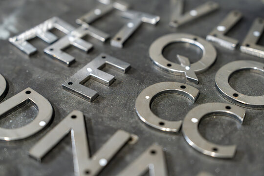 Photographie minimaliste de lettres en m&eacute;tal aux textures industrielles et reflets brillants, isol&eacute;es sur fond neutre pour un rendu moderne et typographique.