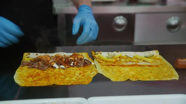 Street chef preparing shawarma wrap with gloved hands, ladle pouring sauce over sliced meat and fries on flatbread, stainless kitchen counter background, closeup of roll assembly and final