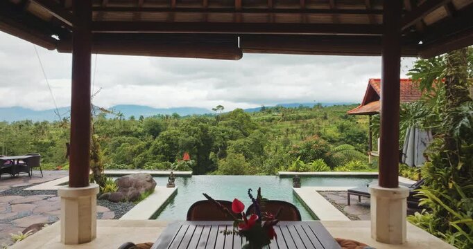 Balinese villa gazebo outdoor dining area with wooden table and chairs, infinity pool overlooking lush green jungle forest and distant mountains under a cloudy sky. Travel, explore Bali. Drone flight