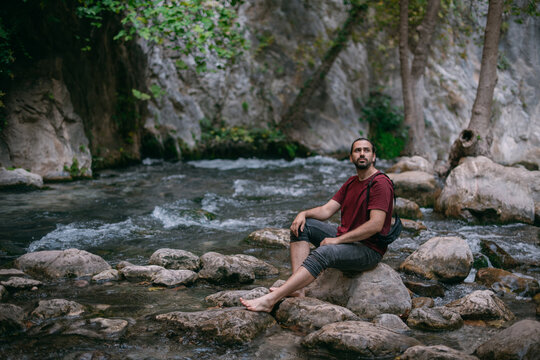 A young male tourist near a clear mountain stream in the rocks overgrown with greenery, on a summer day