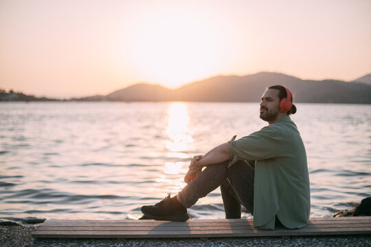 A young man is sitting on the beach with headphones on at sunset by the sea.