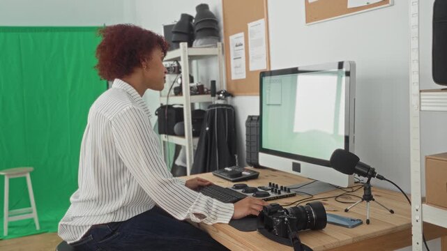 Woman seated at desk typing on keyboard while looking at monitor with camera on table in studio with green screen, microphone and shelves of gear; focused creativity.
