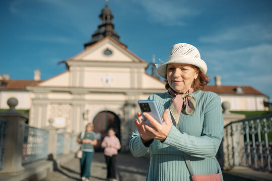 An elderly, contented woman walks with a guide through the park next to an ancient castle. The pensioner examines the historical architecture, looks at a map and reads about the sights on her phone.