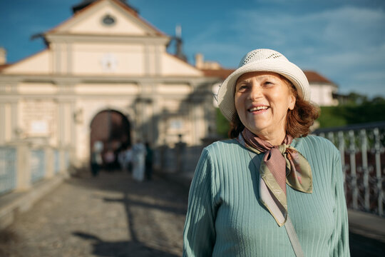 Пожилая довольная женщина гуляет на экскурсии в парке возле старинного замка. Пенсионерка рассматривает историческую архитектуру.