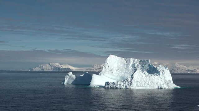 An iceberg is drifting in the ocean water near Greenland. The sunlight reflects off the ice as mountains appear in the background under a blue sky.