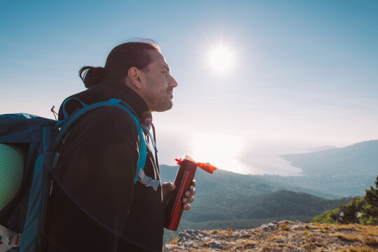 A man hiker drinks tea from a thermos on a mountain top at dawn.